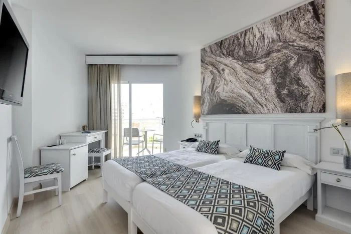 pokój w kolorze białym z łóżkami, biurkiem, obrazem i balkonem